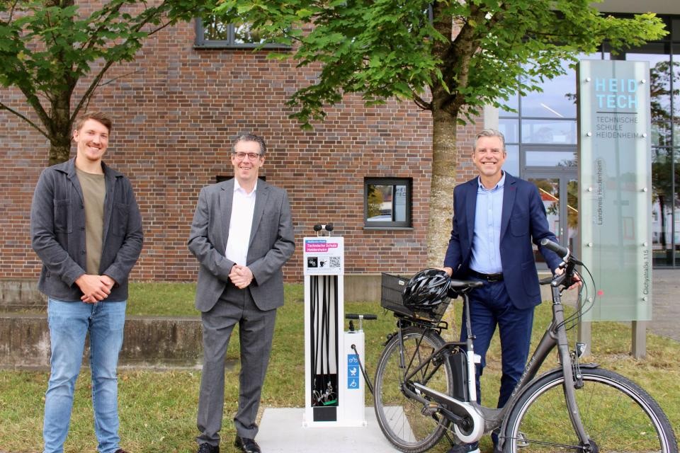 3 Männer und ein Fahrrad stehen vor einer Anlage, mit der man Fahrräder reparieren kann. 